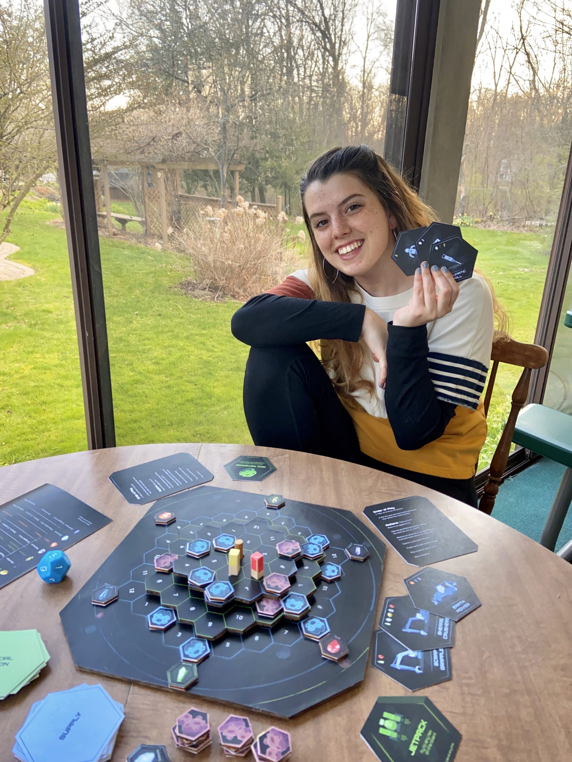 A picture of Kat holding up cards and sitting in front of her board game.